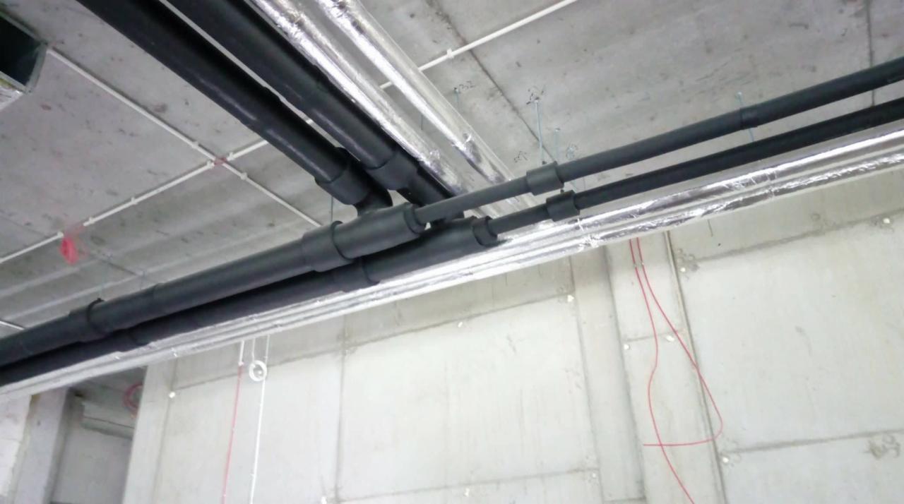 Czarne, zaizolowane rury biegnące pod betonowym sufitem, widoczne łączenia i fragmenty rur owinięte folią aluminiową.