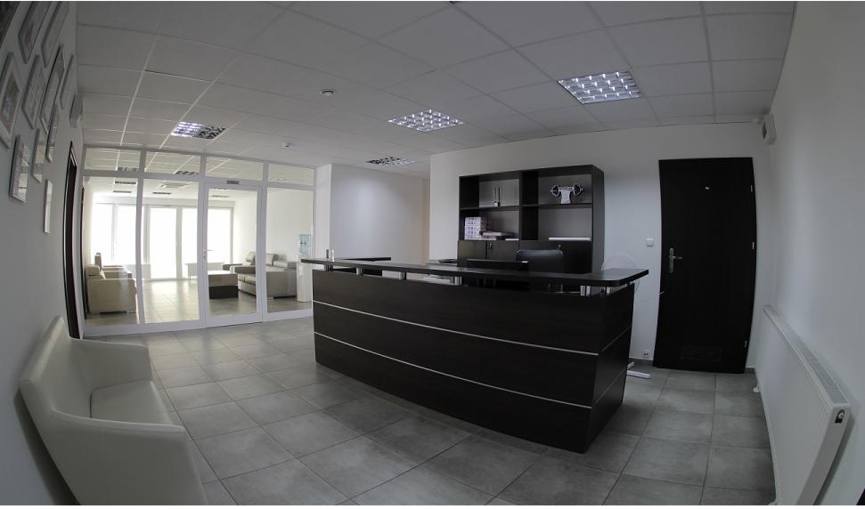 Nowoczesna recepcja biurowa z ciemnym biurkiem recepcyjnym z poziomymi, srebrnymi paskami, szarą podłogą, białymi ścianami i przeszklonym wejściem do poczekalni z jasnymi sofami.