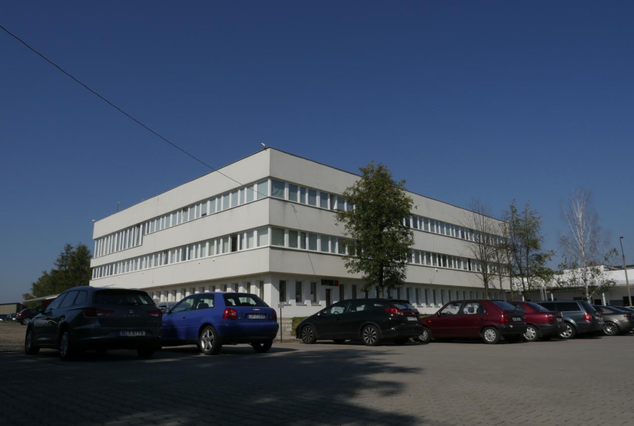 Biały, trzypiętrowy budynek magazynowy w Opolu z rzędem zaparkowanych samochodów osobowych przed nim.