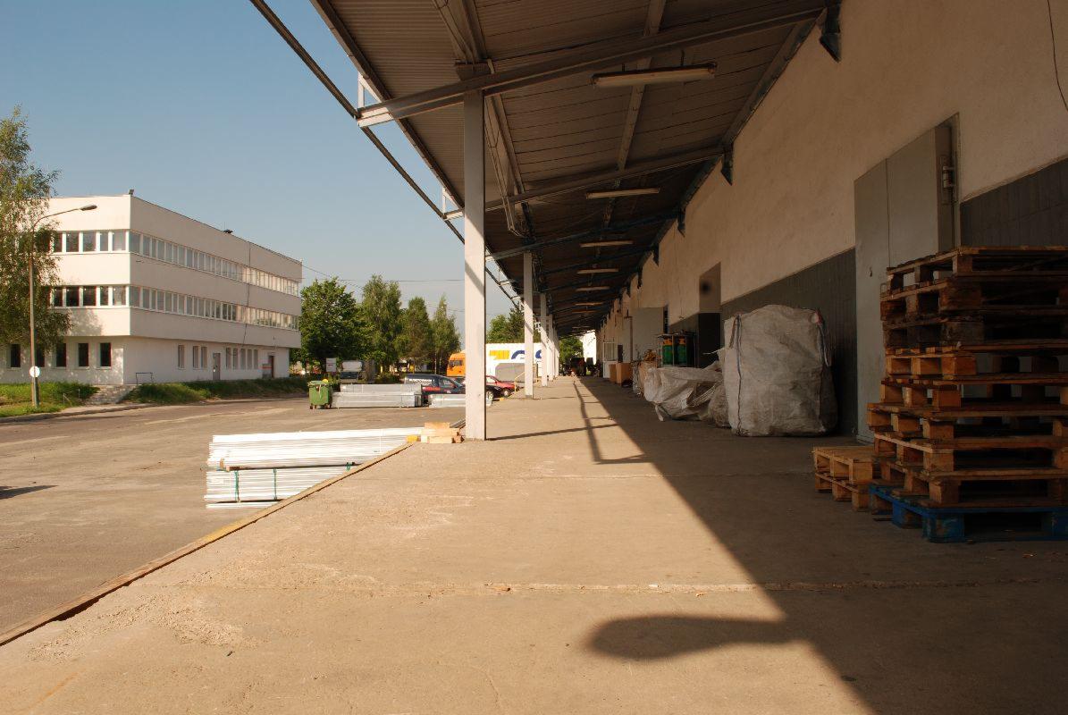 Zadaszony plac magazynowy z paletami, workami i stertą metalowych profili, budynek biurowy w tle.