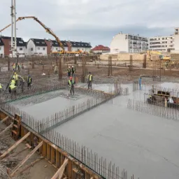 Betonowanie fundamentów budynku wielorodzinnego: ekipa w żółtych kamizelkach rozprowadza świeży beton na zbrojeniu, w tle pompa do betonu i budynki mieszkalne.