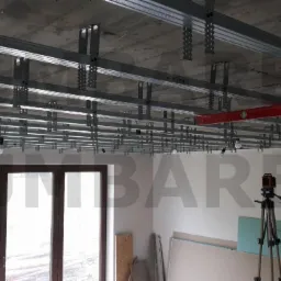 Montaż metalowej konstrukcji sufitu podwieszanego z płyt kartonowo-gipsowych, widoczny laser krzyżowy na statywie, płyty g-k oparte o ścianę.