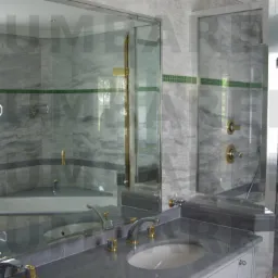 Elegancka łazienka wykończona marmurem z kabiną prysznicową ze złotymi okuciami i umywalką wpuszczaną w szary blat.