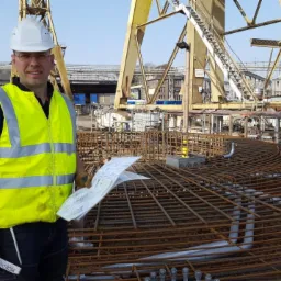 Pracownik w kamizelce odblaskowej i kasku trzymający plany na tle zbrojenia fundamentu pod budowlą przemysłową, z widocznym dźwigiem i innymi pracownikami w oddali.