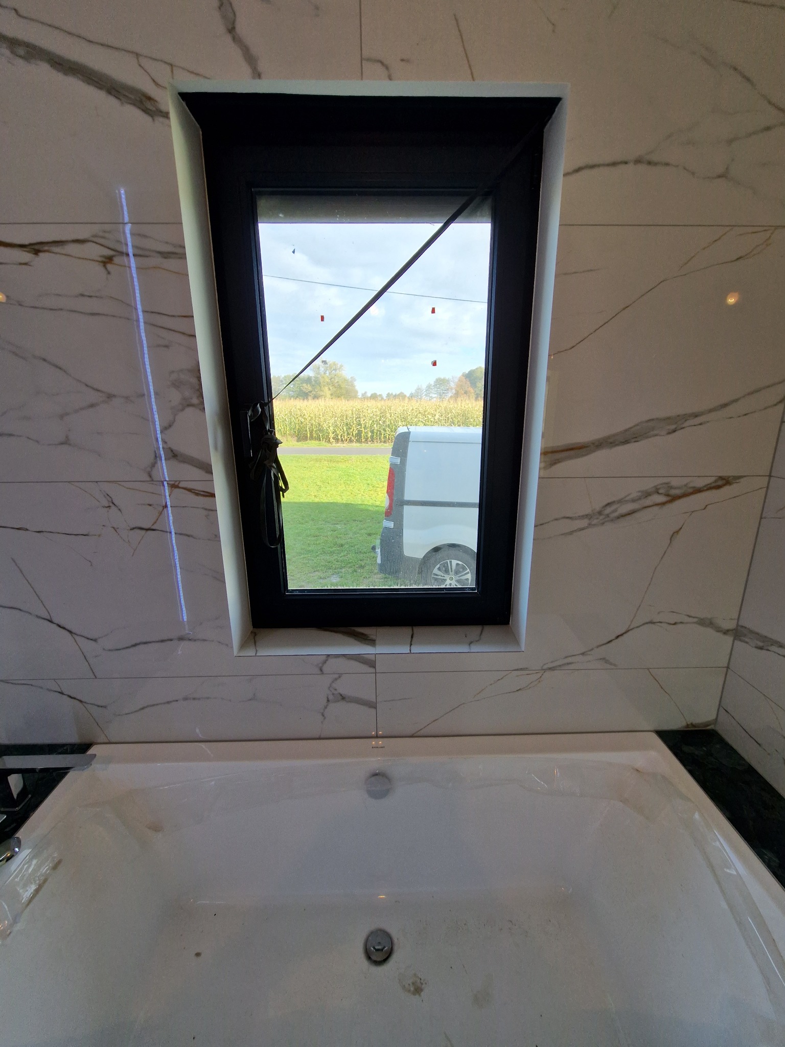 Łazienka z wanną, okno z widokiem na pole kukurydzy i zaparkowany biały samochód dostawczy, marmurowe płytki na ścianach.