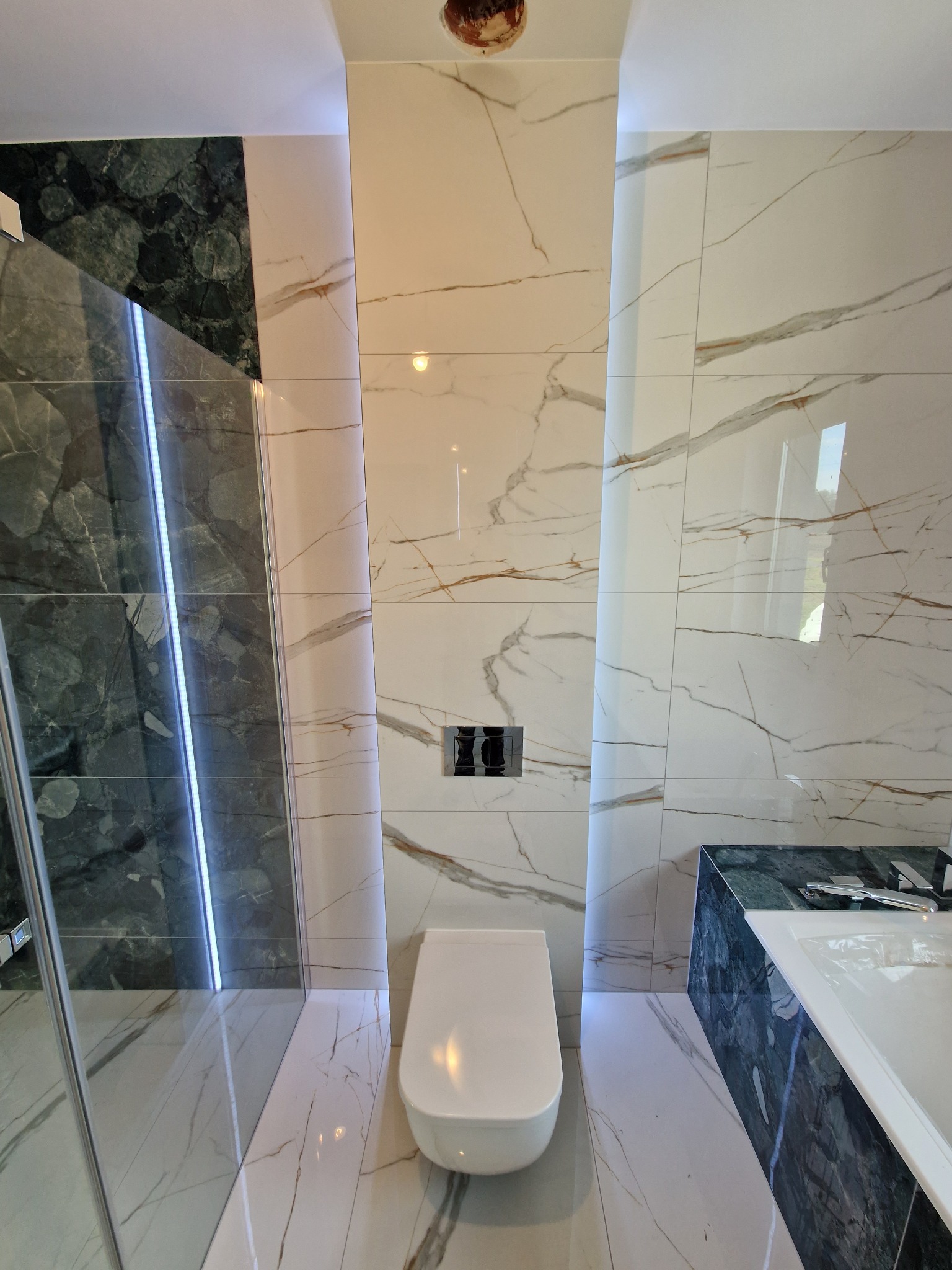 Nowoczesna łazienka z białymi płytkami imitującymi marmur z brązowymi żyłami, podświetleniem LED i elementami z ciemnozielonego marmuru, widoczna toaleta podwieszana i fragment kabiny prysznicowej.