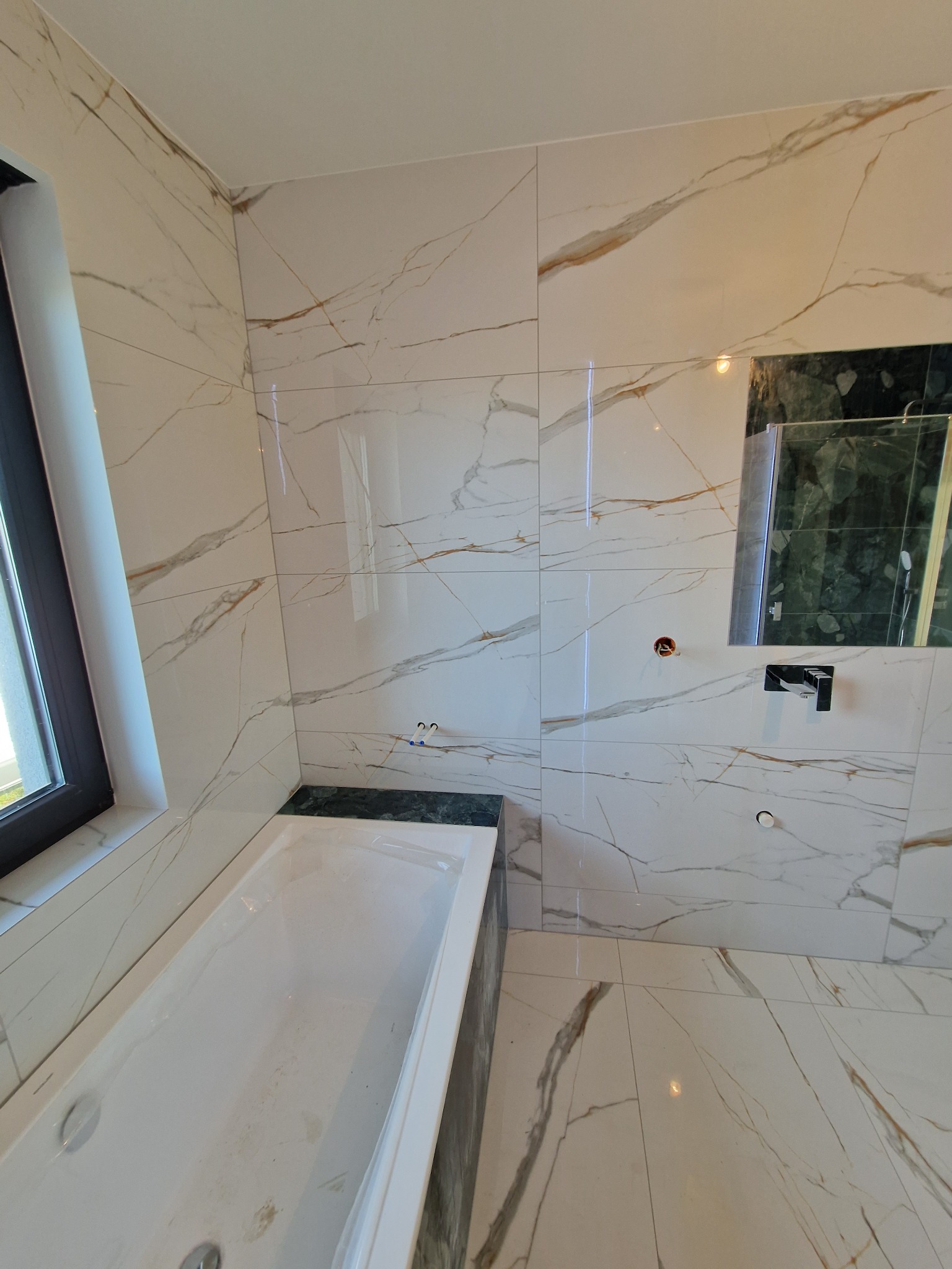 Wnętrze łazienki po remoncie z wanną obudowaną ciemnym kamieniem, wykończone płytkami imitującymi marmur z delikatnymi złotymi żyłkami, widoczne otwory na baterie, jeszcze nie zamontowane.