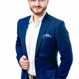 Portret uśmiechniętego mężczyzny w okularach, w granatowym garniturze i białej koszuli, z ozdobną poszetką w kieszonce, pozującego na jasnym tle.