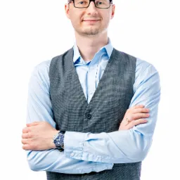 Mężczyzna w okularach, błękitnej koszuli i szarej kamizelce, z założonymi rękami, na białym tle.