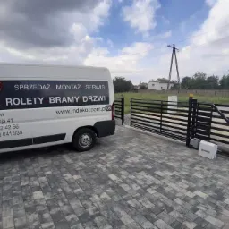 Czarna brama przesuwna z automatyką na posesji, obok zaparkowany biały bus firmy Indekor z reklamą okien, rolet, bram i drzwi.