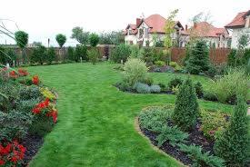 Rozległy, zadbany trawnik z równo przystrzyżoną trawą, otoczony rabatami kwiatowymi i krzewami ozdobnymi. W tle widoczny dom z czerwoną dachówką i ogrodzenie.