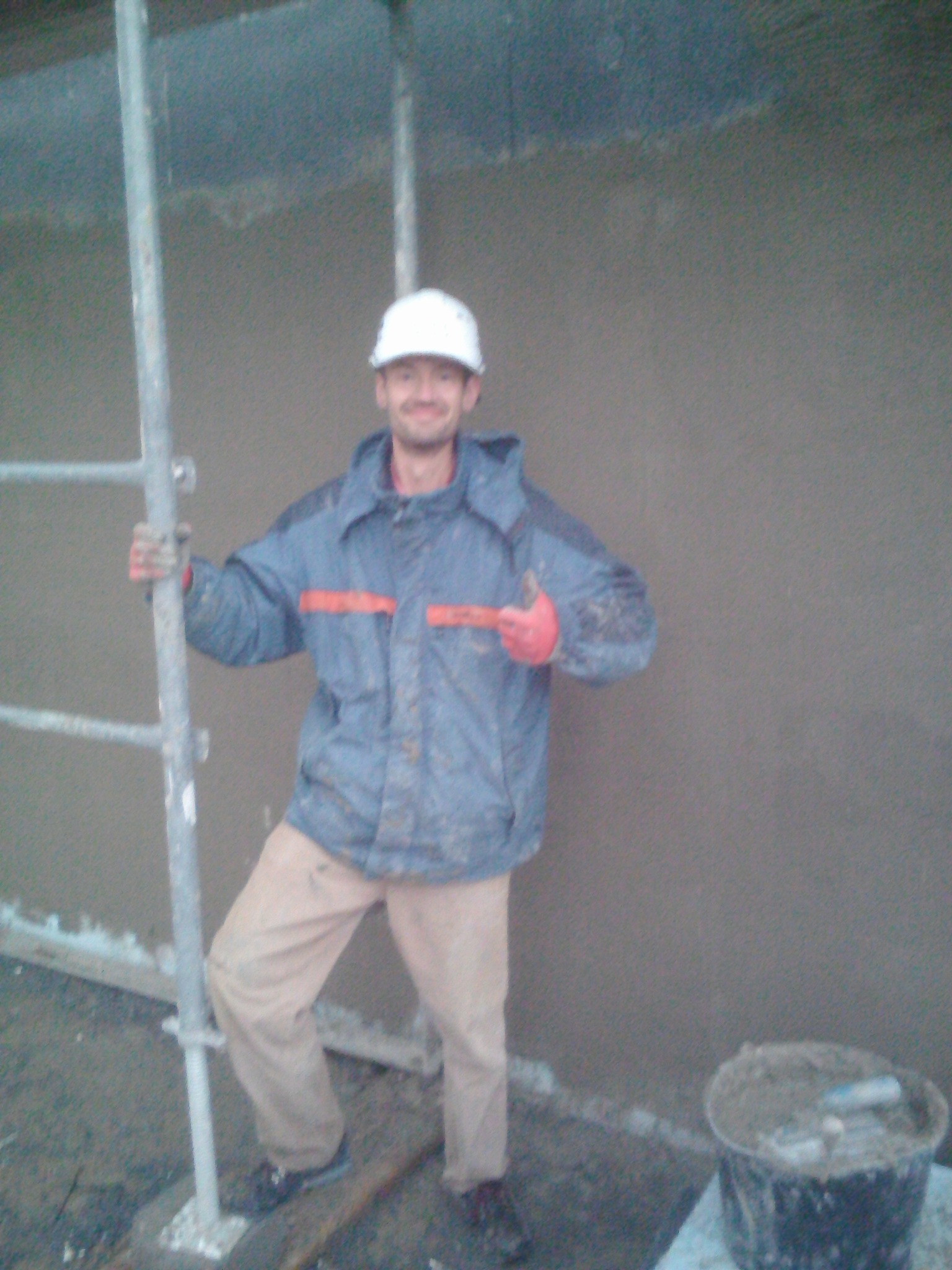 Mężczyzna w kasku i roboczym ubraniu stoi przy rusztowaniu na tle świeżo otynkowanej ściany, obok stoi wiadro z zaprawą.