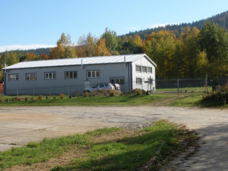 Szara hala z blachy trapezowej z płaskim dachem na tle lasu jesienią, widoczna z zewnątrz, przed budynkiem zaparkowany samochód.