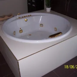 Biała wanna z hydromasażem, obudowana jasnymi płytkami z delikatnym wzorem drewna i złotymi akcentami, na tle ciemnobrązowych płytek podłogowych.