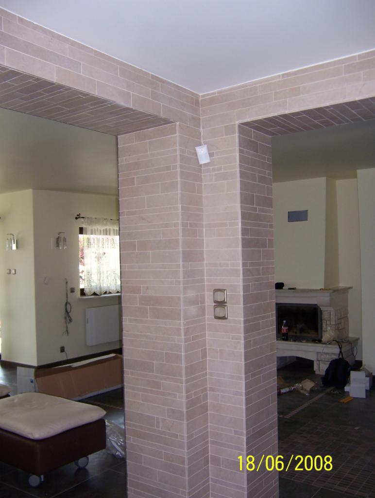 Wykończona kolumna i fragment ściany z beżowych, prostokątnych płytek. Na podłodze ciemne płytki. W tle widoczny kominek i fragmenty wyposażenia wnętrza.