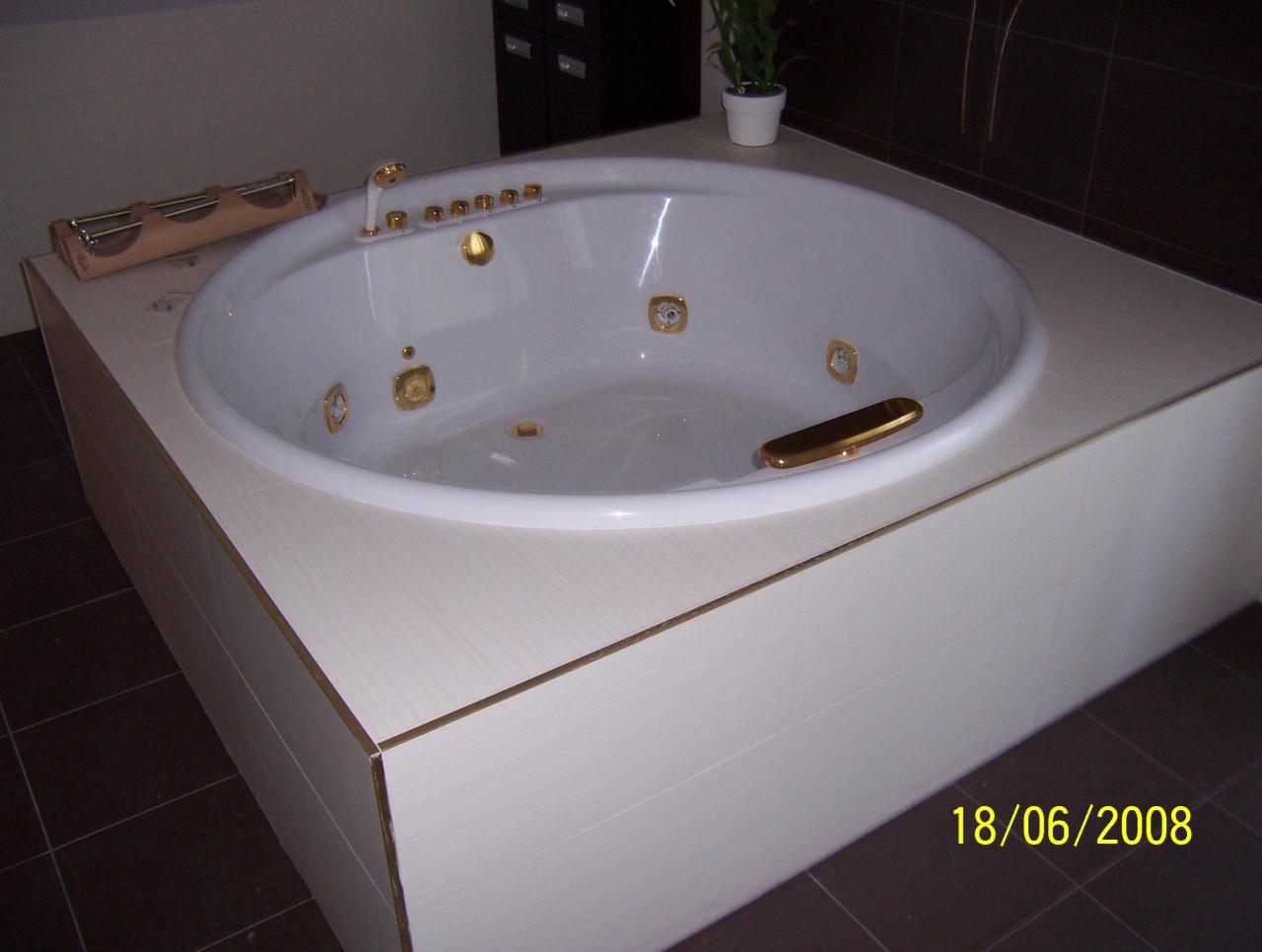 Biała wanna z hydromasażem, obudowana jasnymi płytkami z delikatnym wzorem drewna i złotymi akcentami, na tle ciemnobrązowych płytek podłogowych.