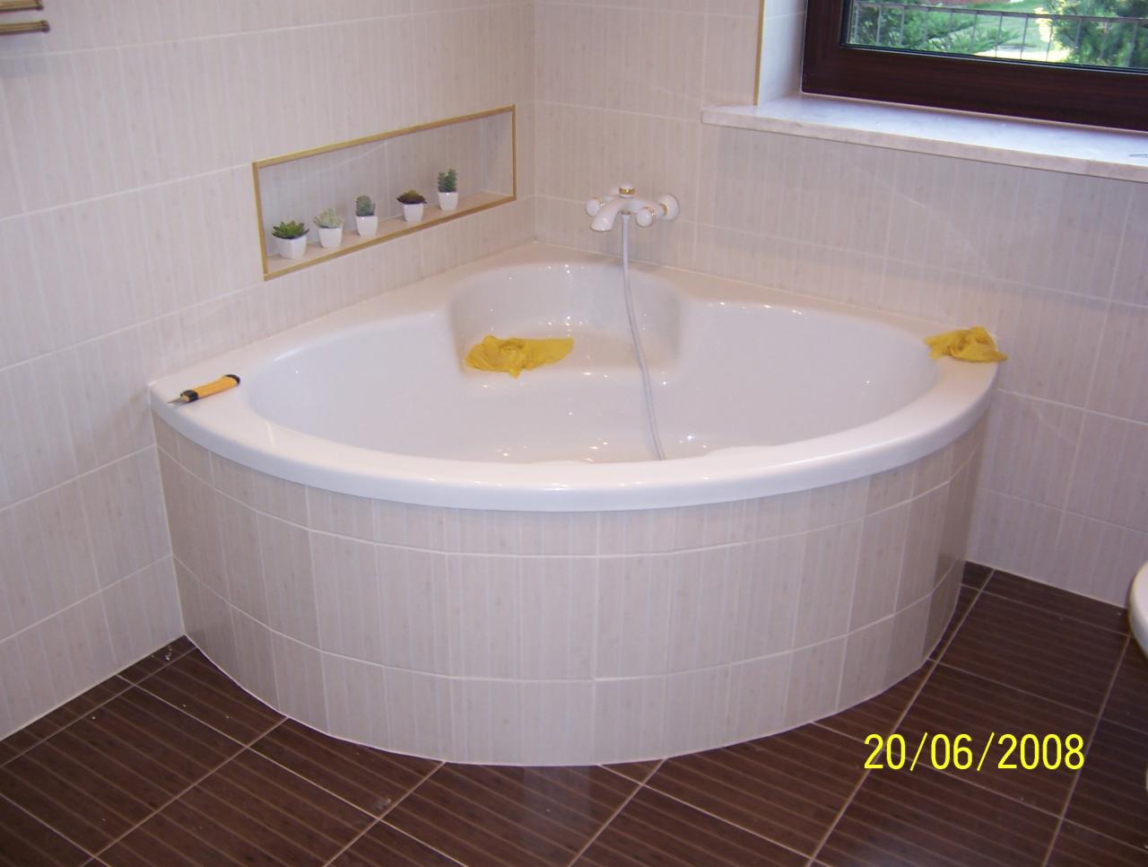 Narożna wanna obudowana beżowymi płytkami, z żółtymi ręcznikami i wnęką z sukulentami, na tle brązowej podłogi z płytek. Widoczny fragment okna.