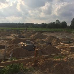 DNV Realiz LTD Sp. Z o.o. S.k - Wykop pod fundamenty domu jednorodzinnego z widocznymi szalunkami, zbrojeniami i pracownikami na placu budowy, w tle las i pochmurne niebo.