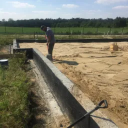 Pracownik wyrównuje teren wewnątrz fundamentów, częściowo zabezpieczonych czarną izolacją, widoczna łopata i wiadro, w tle zieleń i błękitne niebo.