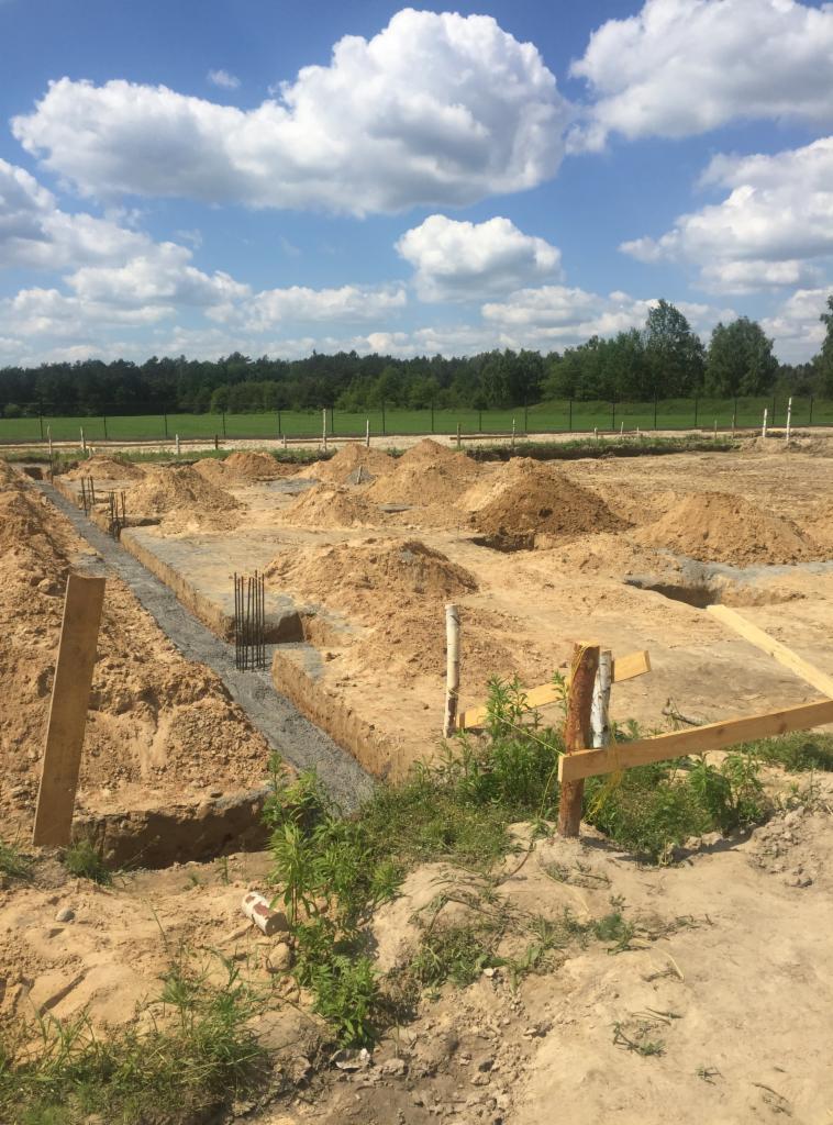Wykopane fundamenty z częściowo zalanym betonem i wystającymi prętami zbrojeniowymi, otoczone piaskiem i ziemią, z widocznym ogrodzeniem w tle oraz błękitnym niebem z białymi chmurami.