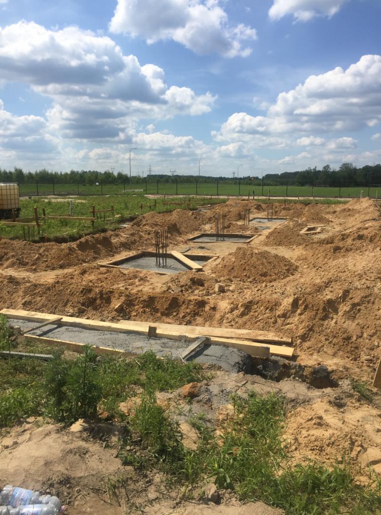 Przygotowanie fundamentów pod budynek: zalane betonem ławy fundamentowe z wystającymi prętami zbrojeniowymi, drewniane szalunki i wykopana ziemia na placu budowy w słoneczny dzień.