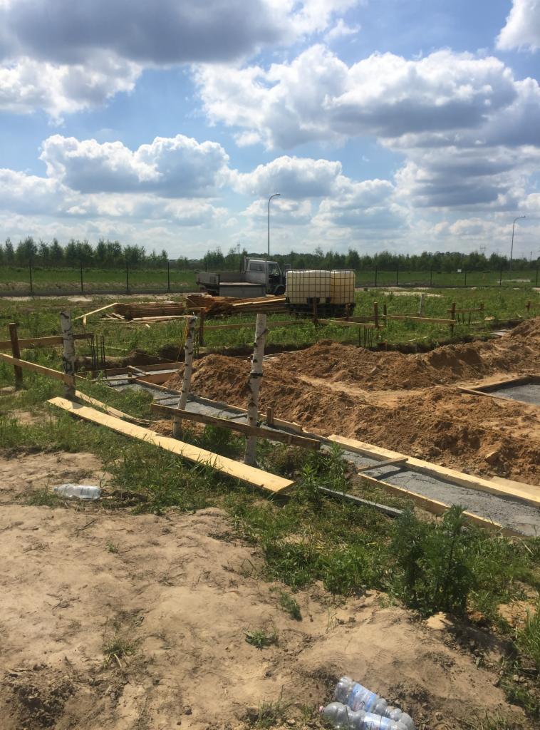 Przygotowanie terenu pod fundamenty: wykopane rowy z betonową wylewką, drewniane szalunki, sterta ziemi, w tle ciężarówka z beczką na wodę i słupy elektryczne.