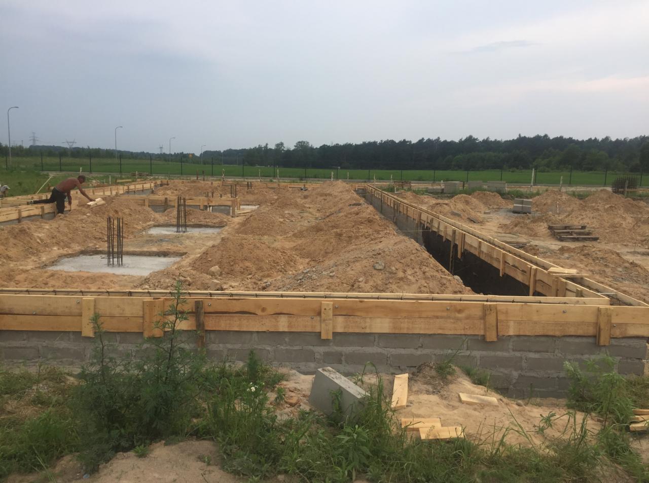 Fundamenty domu jednorodzinnego w trakcie budowy: szalunki z desek, betonowe wylewki z wystającymi prętami zbrojeniowymi, pracownik w tle.