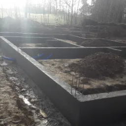 Wylane fundamenty domu jednorodzinnego z wystającymi prętami zbrojeniowymi i niebieskimi rurami instalacyjnymi w ziemi, w tle drzewa i mgła.