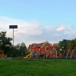 Plac zabaw oświetlony lampami solarnymi, w tle budynek z czerwoną dachówką.