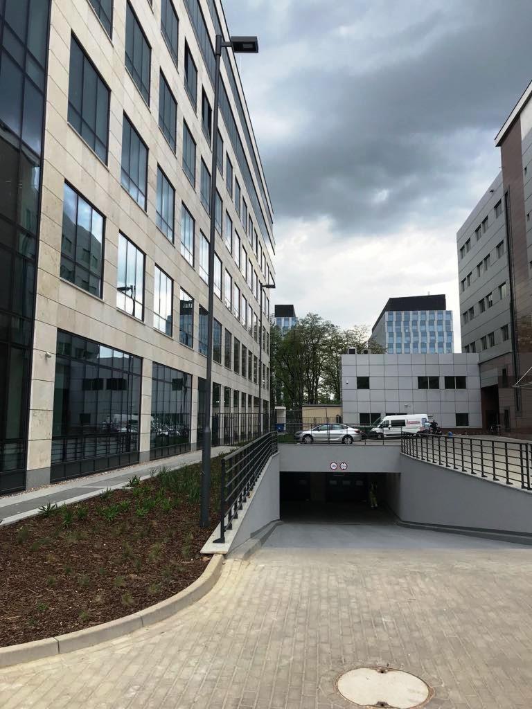 Nowoczesny budynek biurowy z oświetleniem zewnętrznym w postaci słupa oświetleniowego, widok na wjazd do parkingu podziemnego i pochmurne niebo.