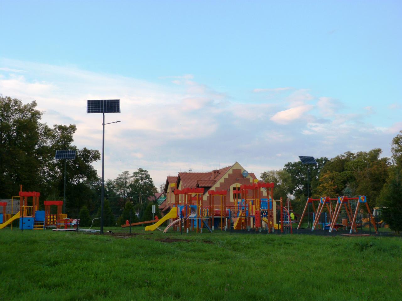 Plac zabaw oświetlony lampami solarnymi, w tle budynek z czerwoną dachówką.