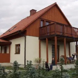 Gotowy dom z drewnianą elewacją, balkonem i czerwoną dachówką, otoczony iglakami, z widoczną kobietą siedzącą na ganku z psem i dzieckiem na huśtawce.