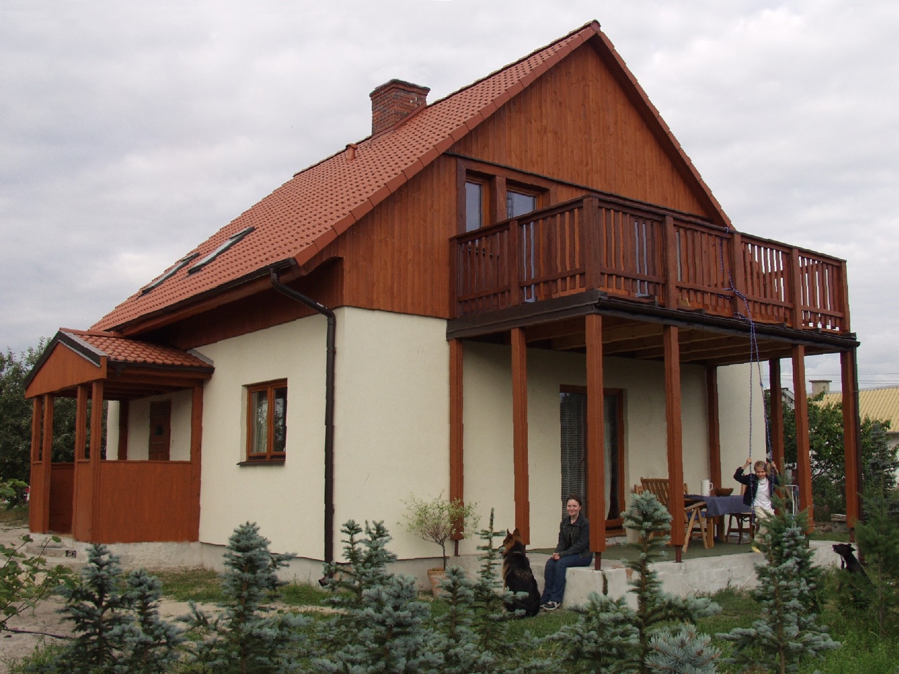 Gotowy dom z drewnianą elewacją, balkonem i czerwoną dachówką, otoczony iglakami, z widoczną kobietą siedzącą na ganku z psem i dzieckiem na huśtawce.