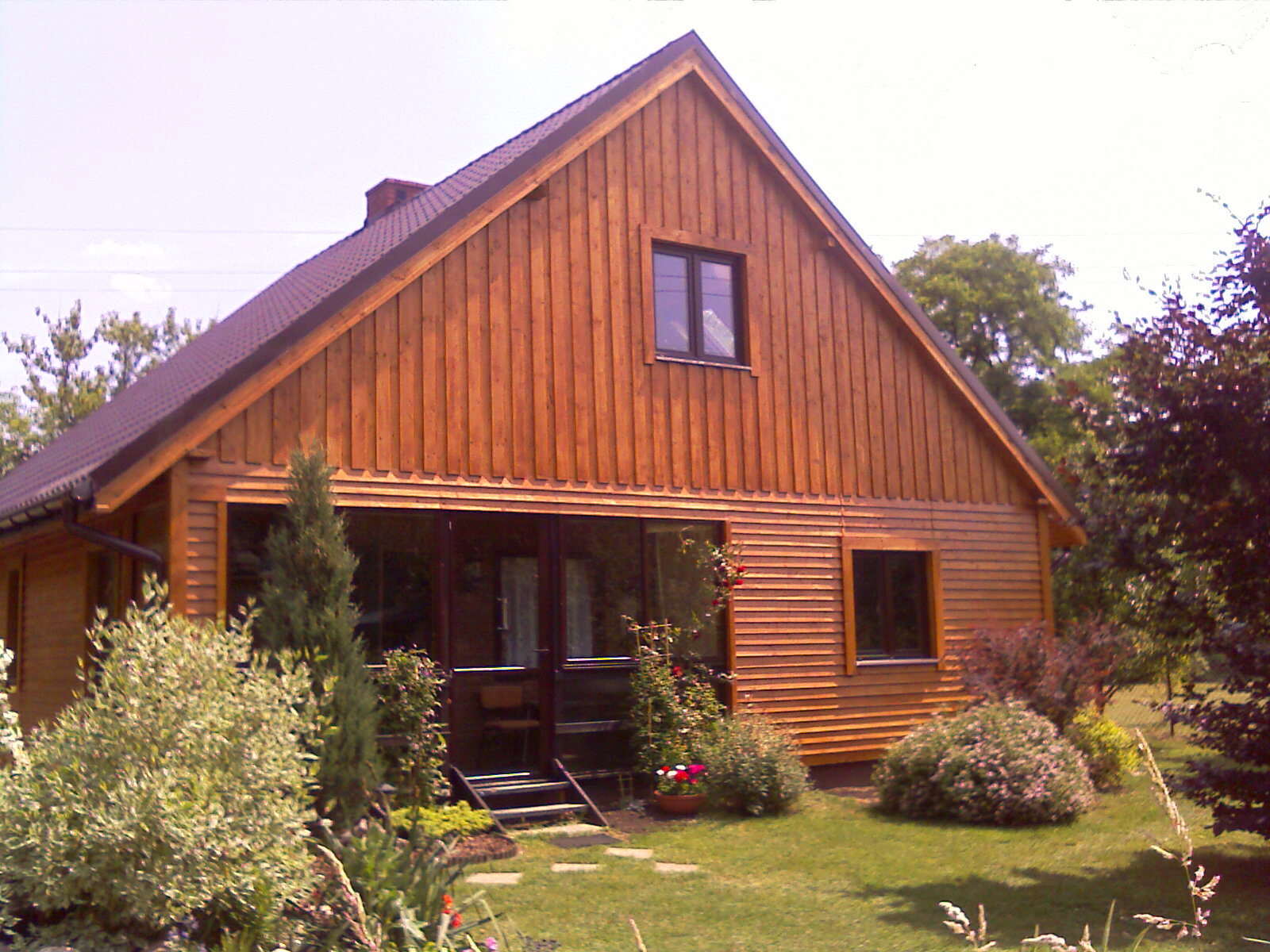 Gotowy drewniany dom z brązową dachówką, widoczne duże okno i drzwi wejściowe, otoczony zielenią ogrodu.