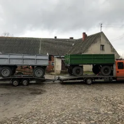 Pomarańczowa laweta Renault Master transportuje dwa traktory rolnicze z przyczepami na tle wiejskiego krajobrazu z budynkami gospodarczymi.