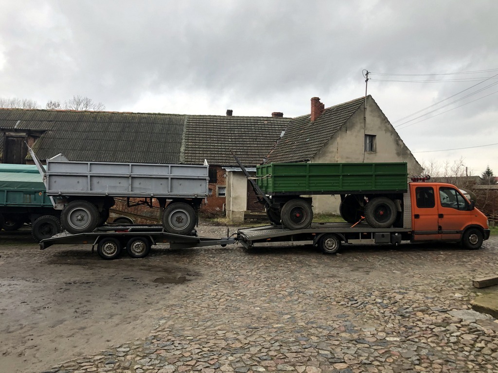 Pomarańczowa laweta Renault Master transportuje dwa traktory rolnicze z przyczepami na tle wiejskiego krajobrazu z budynkami gospodarczymi.