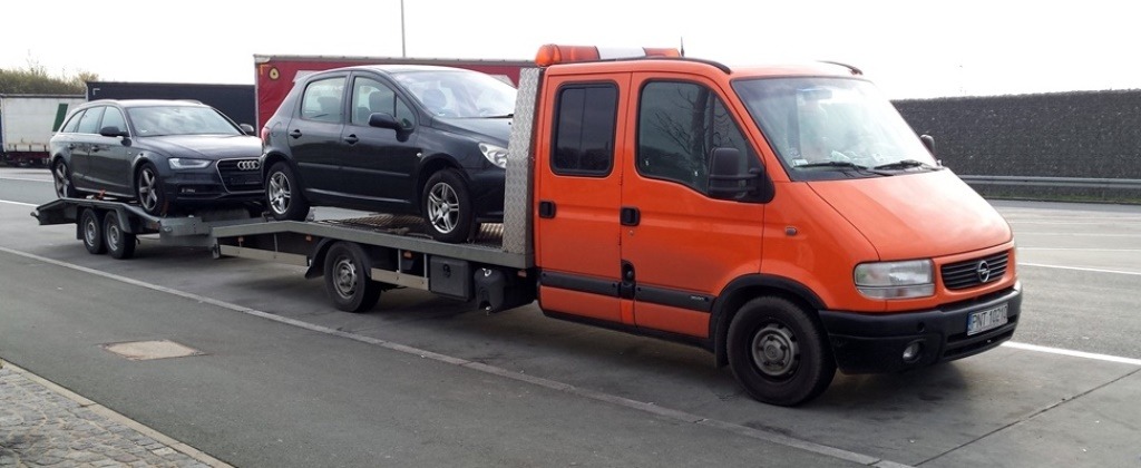 Pomarańczowa laweta z dwoma samochodami osobowymi na platformie, Audi kombi i Peugeot 307, zaparkowana na poboczu drogi w pochmurny dzień.