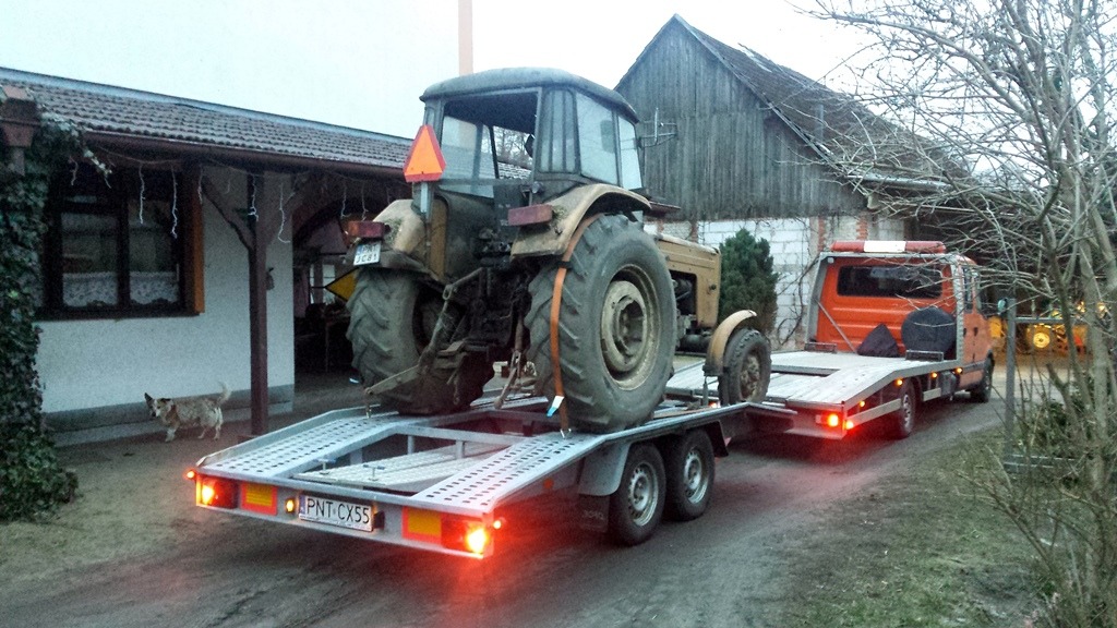 Używany traktor Ursus na niskopodwoziowej przyczepie lawety, transportowany na posesji z budynkiem mieszkalnym i gospodarczym w tle, mały pies obserwuje.