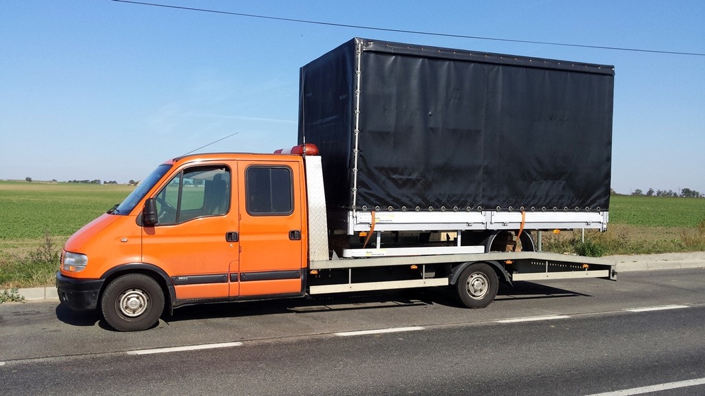 Pomarańczowa laweta z czarną plandeką na platformie, stojąca na poboczu drogi w słoneczny dzień.