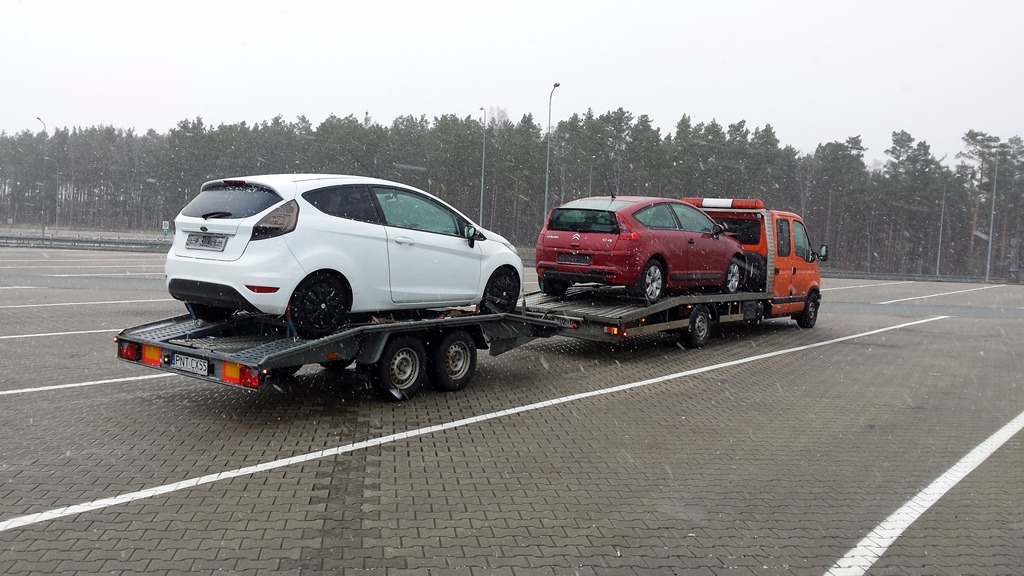 Pomarańczowa laweta z dwoma samochodami (białym Fordem Fiesta i czerwonym Citroenem C4) na parkingu w pochmurny dzień. W tle widoczny las.