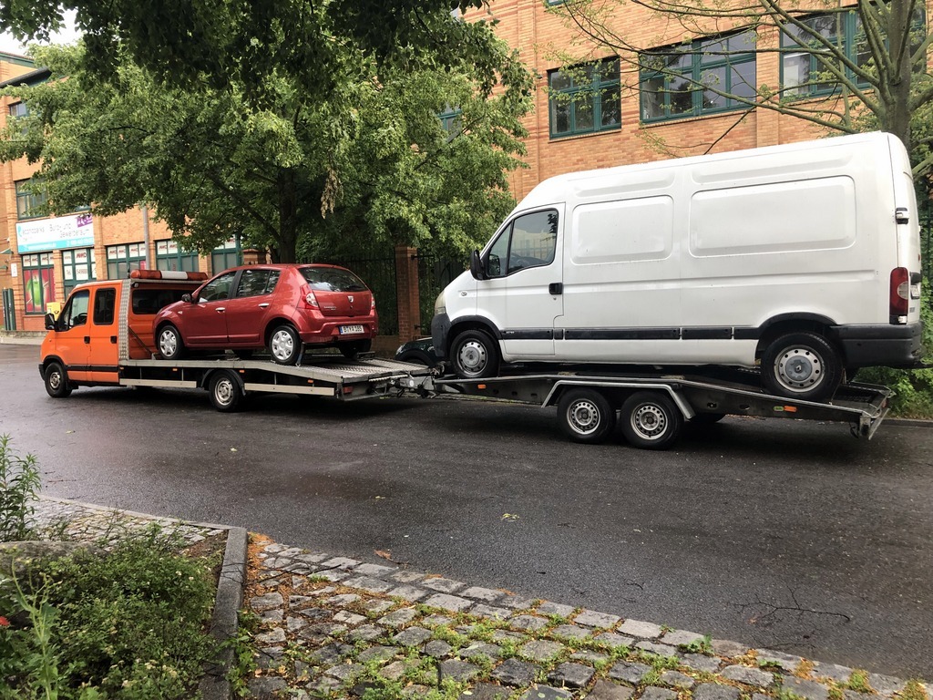 Pomarańczowa laweta z czerwonym samochodem osobowym, za nią biały bus na przyczepie, transport na mokrej ulicy w tle budynek z czerwonej cegły i drzewa.
