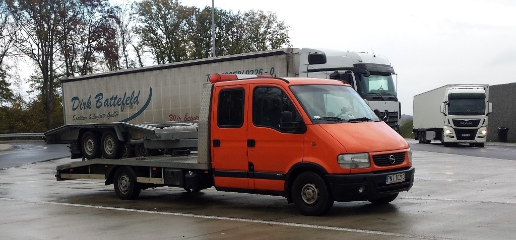 Pomarańczowa laweta na parkingu, z przyczepą transportującą podwozie naczepy, w tle inne samochody ciężarowe.
