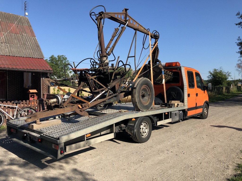 Pomarańczowa laweta przewozi starą, rdzewiejącą konstrukcję metalową z elementami przypominającymi dźwig, zabezpieczoną pasami transportowymi, w tle wiejski krajobraz z domem i rowerem.