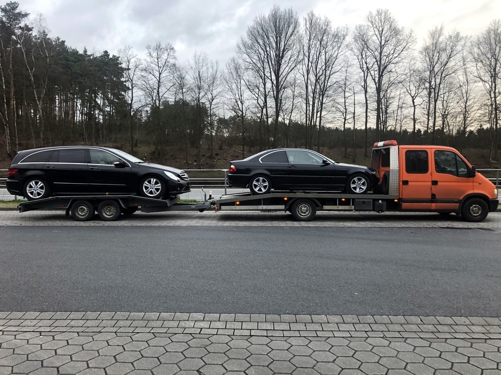 Pomarańczowa laweta przewozi dwa czarne samochody osobowe na dwuosiowych przyczepach, widoczne drzewa w tle.