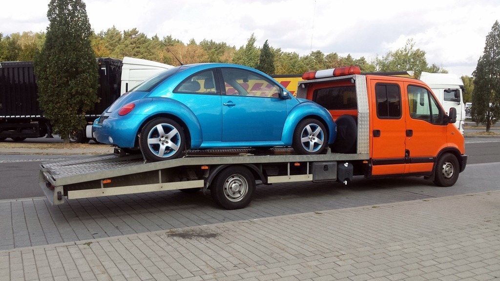 Niebieski Volkswagen Beetle na platformie lawety pomalowanej na pomarańczowo-srebrny kolor, zaparkowanej na brukowanym placu, z ciężarówkami i drzewami w tle.