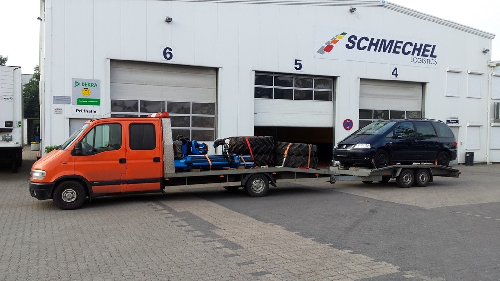 Pomarańczowa laweta z niebieskim urządzeniem i oponami na platformie, ciągnąca przyczepę z ciemnym minivanem na tle budynku firmy transportowej Schmechel Logistics.