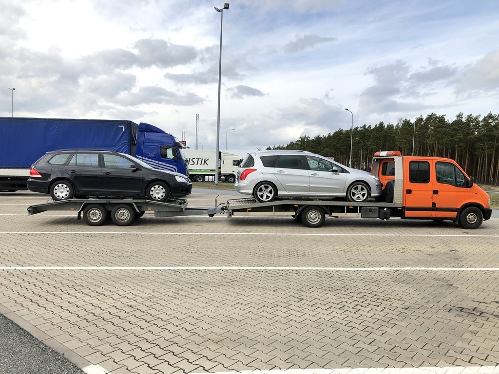 Pomarańczowa laweta z dwoma samochodami osobowymi (czarnym kombi i srebrnym hatchbackiem) na przyczepach, zaparkowana na parkingu z kostki brukowej, w tle inne pojazdy ciężarowe i las