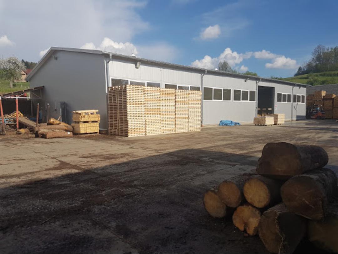 Składowisko drewna z ułożonymi paletami, pociętymi kłodami i budynkiem magazynowym w tle, widoczne wózki widłowe i zieleń na horyzoncie.