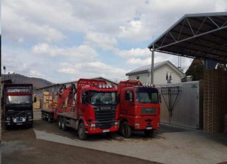 Dwa czerwone samochody ciężarowe Scania i MAN oraz czarny Volvo zaparkowane na placu, jeden z nich załadowany tarcicą budowlaną, w tle budynki i pochmurne niebo.