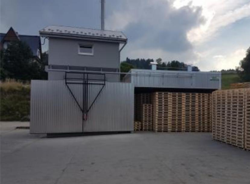 Składowisko palet drewnianych pod zadaszeniem, z metalową konstrukcją i budynkiem w tle, pochmurne niebo.